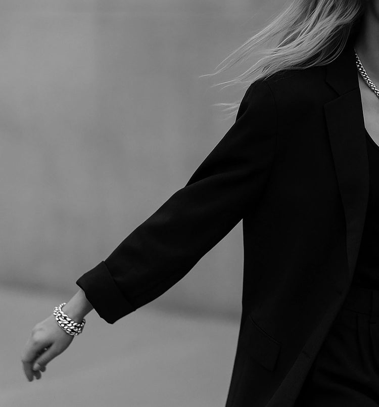 Två chunky silverarmband med stora kedjor bärs av kvinna i svart kostym, hon går på en gata och svänger med armen.
