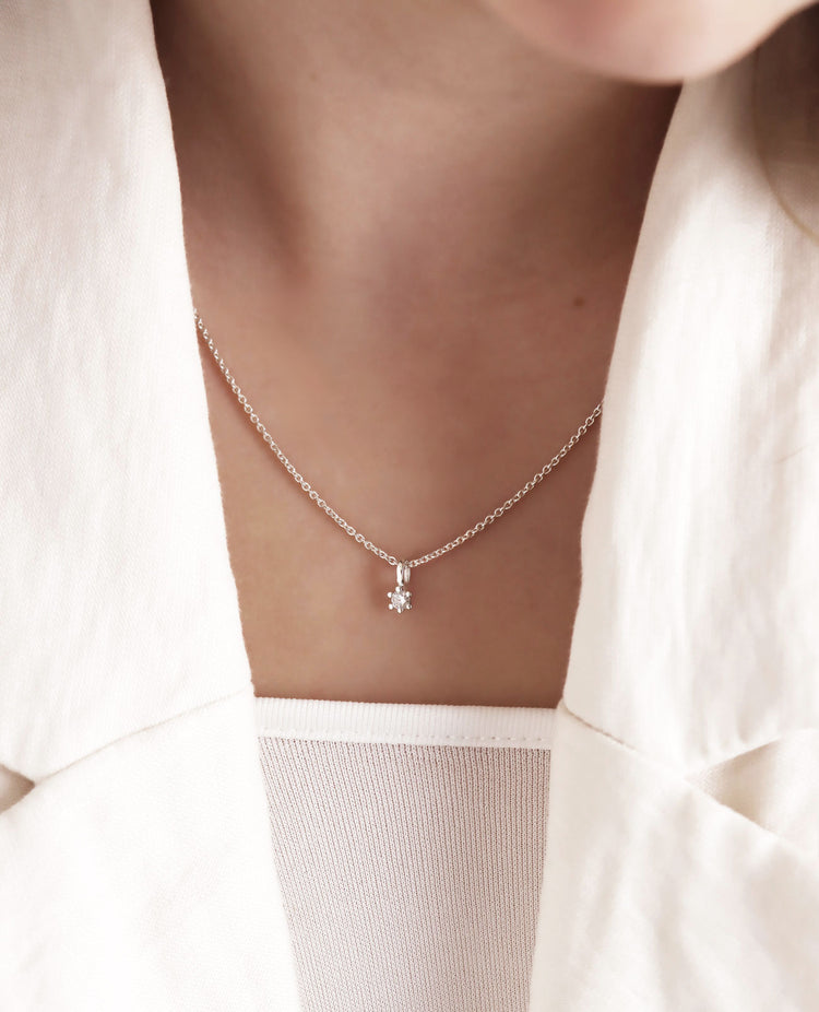 Ett diamanthalsband med ett litet hängsmycke med diamant i silverkedja, bärs av kvinna med vit kavaj och vitt linne.