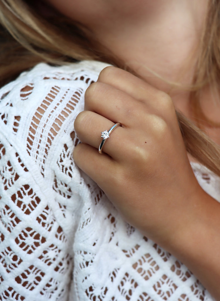 Tjej som bär en diamantring som vigsel- eller förlovningsring med en 0,25 ct diamant. Hon har vit virkad klänning. Diamanten är vit och skiner i solen.