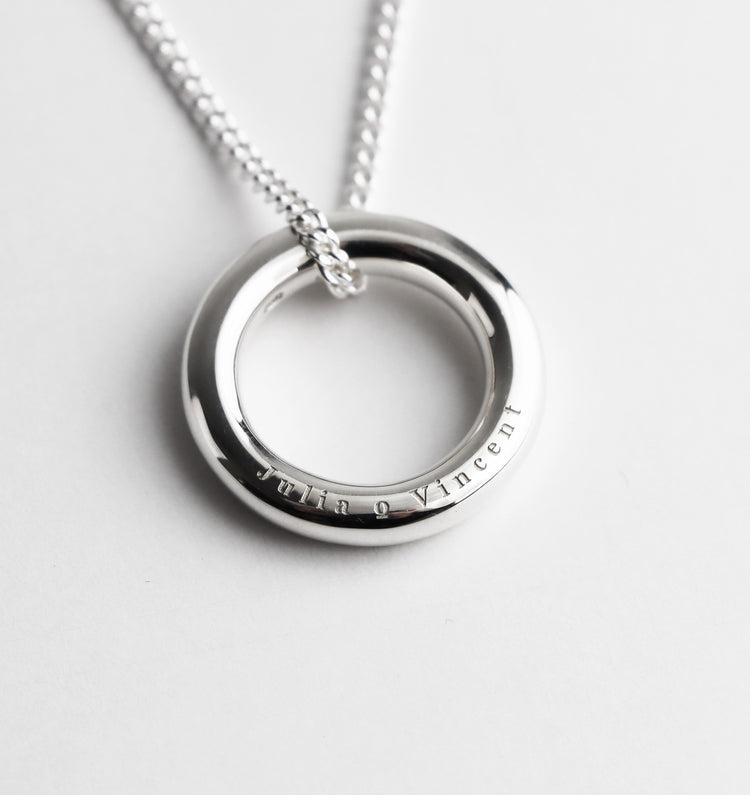 Silverring med två namn graverade på sidan av ringen. Ringen hänger i en halskedja, pansarkedja.