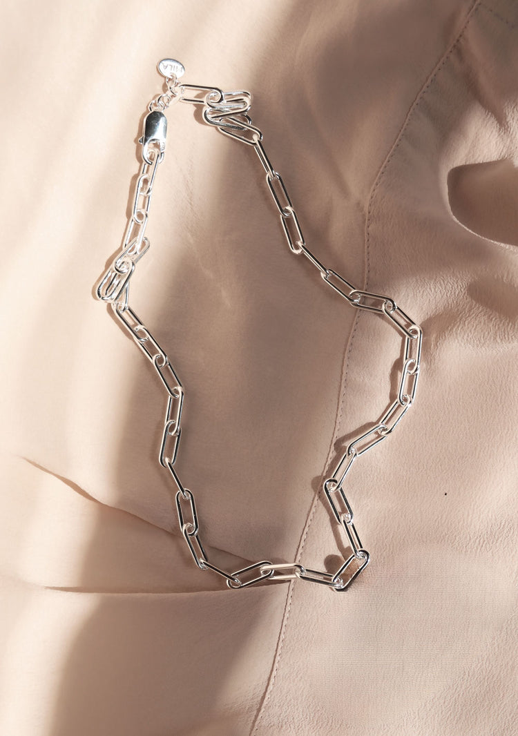 En silverkedja med avlånga länkar ligger på ett rosa tyg, solen strålar in från sidan av bilden.