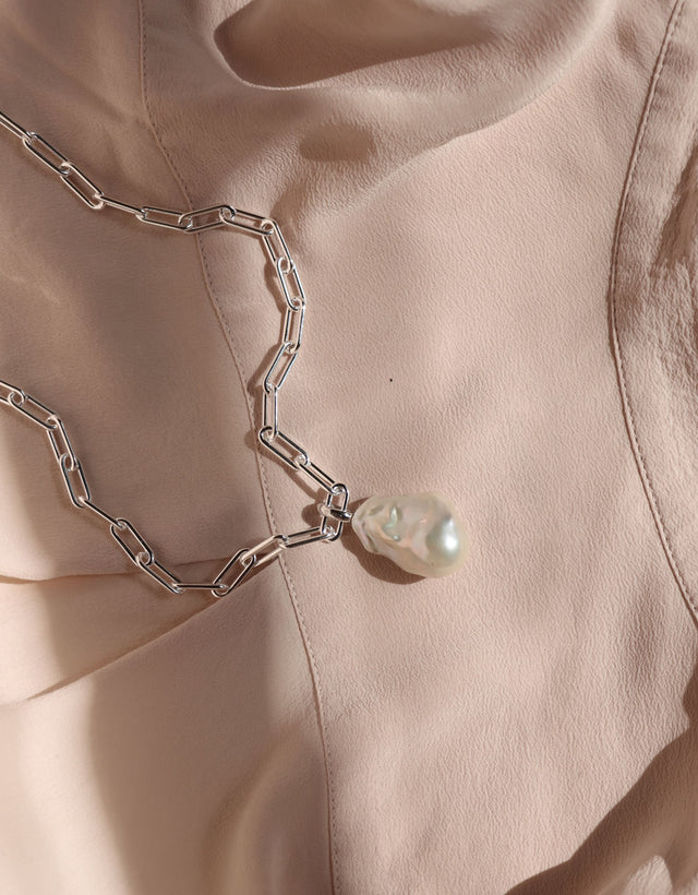 Ett halsband med kedja och stor pärla placerat på ett ljusrosa tyg. 