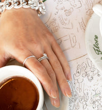 Hand som bär en smal, slät silverring på pekfingret tillsammans med diamantringar i silver. Hon håller i en kaffekopp. 