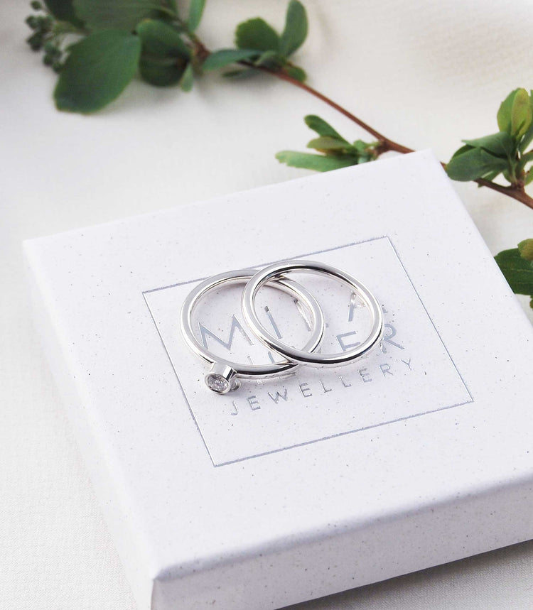 Två silverringar placerade på en ask, den ena ringen är en diamantring med diamant infattad i  bezel.